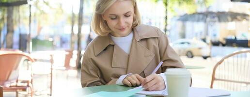 Portrait of carefree, blond cute woman working with documents, drinking takeaway coffee and sitting in cafe outdoors with documents, writing essay, student doing homework photo