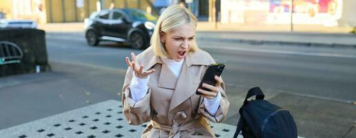 Portrait of angry young woman, shouting at her mobile phone in frustrated, being outraged, sitting on street bench outdoors photo