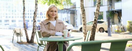 Image of young woman, makes sketches outdoors on street in notebook, drinks coffee and smiles at camera photo