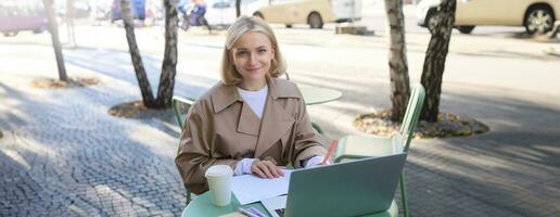 Image of young woman studying outdoors, sitting in cafe, making notes while listening to online lecture using laptop, making notes in notebook, writing photo