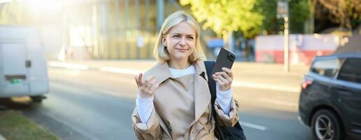 Portrait of confused blonde woman on street, carries backpack, holding smartphone, frowning and looking puzzled, unsure where to go, using mobile map photo