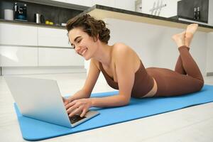 Portrait of beautiful sportswoman, fitness girl watching videos on laptop during workout, follow online pilates videos while exercise at home, wearing activewear in kitchen photo