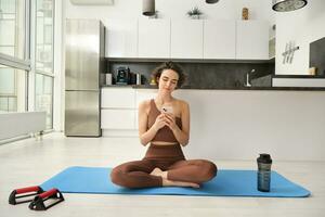 Sport app and wellbeing. Young woman, athlete sitting on floor at home with smartphone, doing yoga, using workout app on mobile phone, doing exercises at home photo