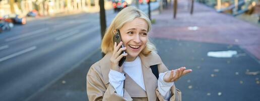 Lifestyle portrait of young blond woman talking on mobile phone, chatting with someone while walking on street, has conversation on smartphone and laughing photo