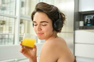 deporte y sano estilo de vida. retrato de contento deportivo chica, aptitud mujer en auriculares, rebobina a hogar, bebidas naranja jugo con contento sonrisa después rutina de ejercicio foto