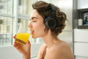 Smiling sportswoman, fitness girl does workout at home, wears headphones, listens music and drinks orange juice after training session photo