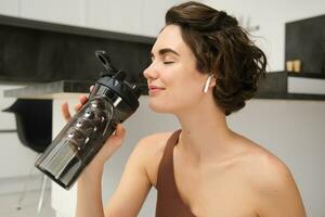 sonriente activo joven mujer en ropa de deporte, se sienta en yoga estera con agua botella, bebidas corsé hidratado durante rutina de ejercicio formación sesión desde hogar, aptitud en el casa en piso foto