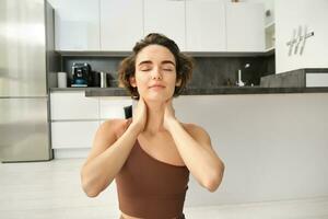 Close up portrait of sportswoman, fitness woman massaging her neck, warm-up her body after workout, doing yoga on rubber mat at home photo