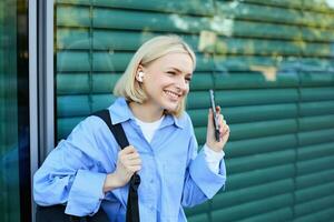 Close up portrait of blond smiling female model, college student in earphones, listening to music on street, holding smartphone, waiting for someone outdoors photo