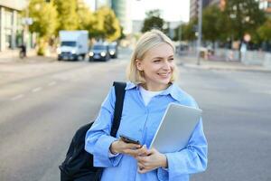 Portrait of blond smiling woman in blue shirt, holding backpack and smartphone, waiting for her ride, order taxi and checking updates on mobile phone, standing on street near road photo