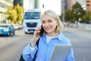 Close up portrait of young woman with laptop, standing on street near busy road in city, talking on mobile phone, answers a call photo