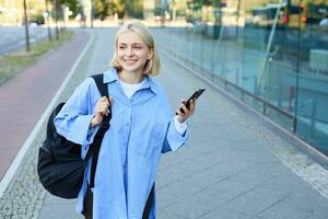 Lifestyle portrait of smiling blond woman with smartphone, walking on street with backpack and looking happy, on her way to college, using mobile phone photo