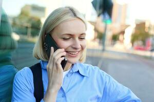 Close up portrait of blond smiling woman, answers a phone call, talking on telephone, standing on street and chatting, looking carefree and happy photo