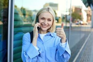Close up portrait of blond smiling woman with backpack, standing on street near office building, showing thumb up in approval, recommending smth good photo