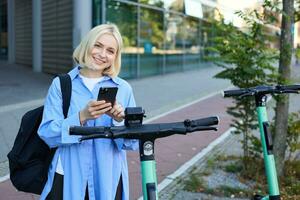 Portrait of smiling young woman unlocks scooter on street, using mobile phone to rent it with smartphone application, riding to university photo