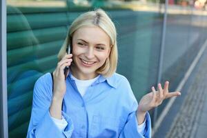 cerca arriba retrato de sonriente rubio mujer, chateando en el teléfono, hablando en móvil teléfono, en pie en calle al aire libre foto