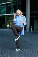 Vertical shot of stylish, happy modern blond girl, student dancing with smartphone near office building, saying yes, making fist pump, celebrating, feeling excitement photo