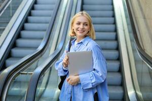 Lifestyle portrait of young woman with laptop, walking away from escalator, heading to tube metro, smiling and looking aside photo