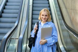 Lifestyle portrait of young blond woman, carries her backpack and laptop, using escalator in city, smiling and looking confident at camera photo