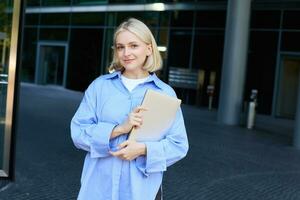 Stylish, modern young woman, student standing on street in campus, heading to lecture with notebook and study material, smiling at camera photo