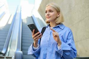 Portrait of beautiful young blond woman in blue shirt, standing near escalator, holding mobile phone, using smartphone, waiting for someone in city centre photo