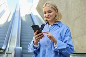 Portrait of young stylish woman, female employee, standing near escalator, using mobile phone, sending message on smartphone, tap screen, smiling and reading on telephone photo