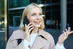Close up portrait of young blond woman, walking on street and talking on mobile phone, chatting with friend on smartphone photo