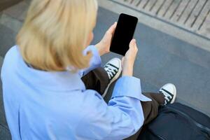 Shot from behind, young woman sitting on stairs on street, holding mobile phone in both hands, screen is dark and blank photo