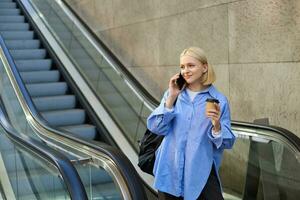 Lifestyle portrait of smiling young woman walking in city, standing on escalator, drinking coffee and chatting on mobile phone photo