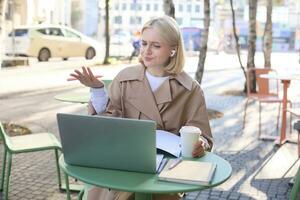 Image of young woman with puzzled face, looking at laptop in wireless headphones, confused while listening to lecture or online speaker, attend web course while sitting in outdoor cafe photo