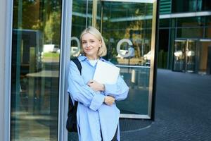 Lifestyle portrait of young woman with laptop, posing near campus, entrance to office building, holding backpack, smiling and looking happy photo