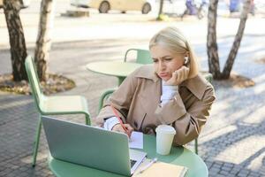 Portrait of blond girl with upset face, listening lecture, attends online course in wireless headphones, sits in outdoor cafe and frowns while makes notes, studying remotely, using laptop photo