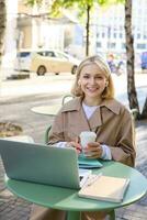 Vertical portrait of smiling blond woman, female student with coffee and laptop, has notebooks on table, studying remotely, working for online project, spending time outdoors on sunny day photo