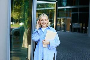 Lifestyle portrait of young woman with laptop, posing near campus, entrance to office building, holding backpack, smiling and looking happy photo