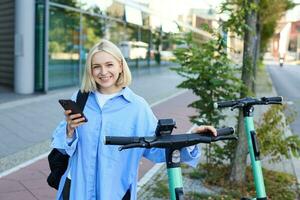 Portrait of young woman, renting a scooter, using mobile phone app to unlock it, using quick ride to get to work, smiling and looking happy photo