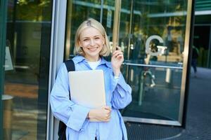 Portrait of carefree young woman, student with laptop, and backpack, leaning on campus building, smiling and laughing, studying and receiving scholarship in university photo