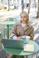 Vertical shot of young blond woman in wireless headphones, making notes, studying online, attend web lecture, using laptop, writing in notebook, looking serious at camera photo