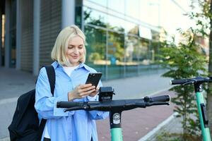 Portrait of young woman, renting a scooter, using mobile phone app to unlock it, using quick ride to get to work, smiling and looking happy photo