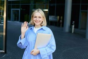 Friendly smiling student, blond girl with notebooks, tutor waving hand at camera, saying hello, standing near office building outdoors photo