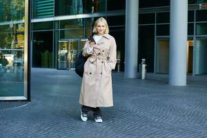 Image of stylish, modern young woman on street, posing in trench coat with backpack, standing and smiling, holding smartphone photo