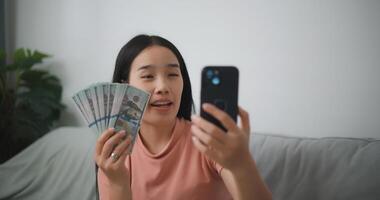 retrato de contento joven asiático mujer sentado en un emocionado estado, participación un en línea teléfono inteligente y dólar cuentas en su mano, ahorra dinero foto