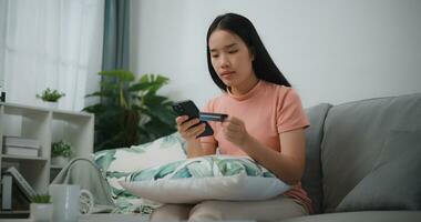 retrato de joven asiático mujer sentado en sofá en vivo habitación, hace en línea bancario pagos mediante el Internet desde banco tarjeta en teléfono inteligente ,compras en línea con crédito tarjeta. foto
