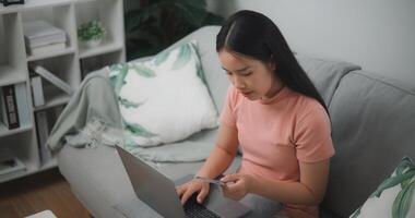 retrato de joven asiático mujer sentado en sofá en vivo habitación, hace en línea bancario pagos mediante el Internet desde banco tarjeta en portátil, compras en línea con crédito tarjeta. foto