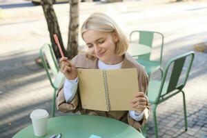 Portrait of young creative woman, doing sketches outdoors, sitting in coffee shop with notebook and pen, drawing art and smiling photo