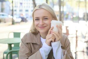 Close up of cheerful blond woman, holding cup of coffee, drinking tea in street outdoor cafe, smiling with happy face expression photo