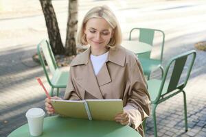 Portrait of young creative woman, doing sketches outdoors, sitting in coffee shop with notebook and pen, drawing art and smiling photo