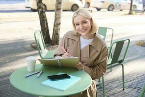 Portrait of beautiful blond woman, sitting in outdoor coffee shop, drawing in cafe in notebook, making sketches outside on street photo