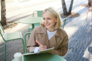 Image of young smiling woman, sitting in outdoor cafe, doing sketches, drawing art on street, spending time on coffee shop and creating art photo