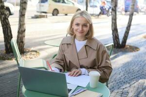 Image of young woman studying outdoors, sitting in cafe, making notes while listening to online lecture using laptop, making notes in notebook, writing photo