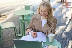 Image of young woman studying outdoors, sitting in cafe, making notes while listening to online lecture using laptop, making notes in notebook, writing photo